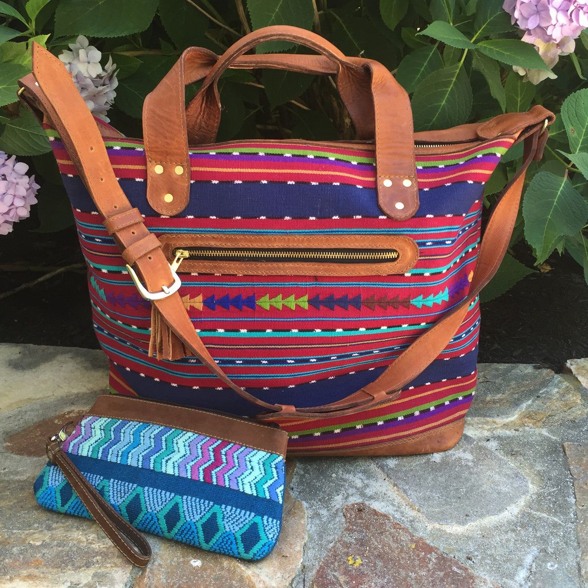 The Weekender Travel Bag with Randa Accent alongside a blue clutch.  Sitting on stone ground in front of a plant.