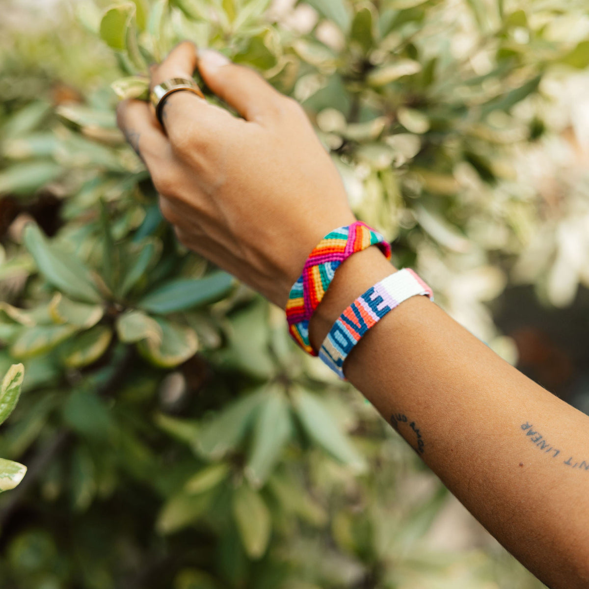 A close-up of a person's hand and forearm wearing colorful woven friendship bracelets, including the LOVE Color Block Bracelet, against a soft-focus background of green leaves.