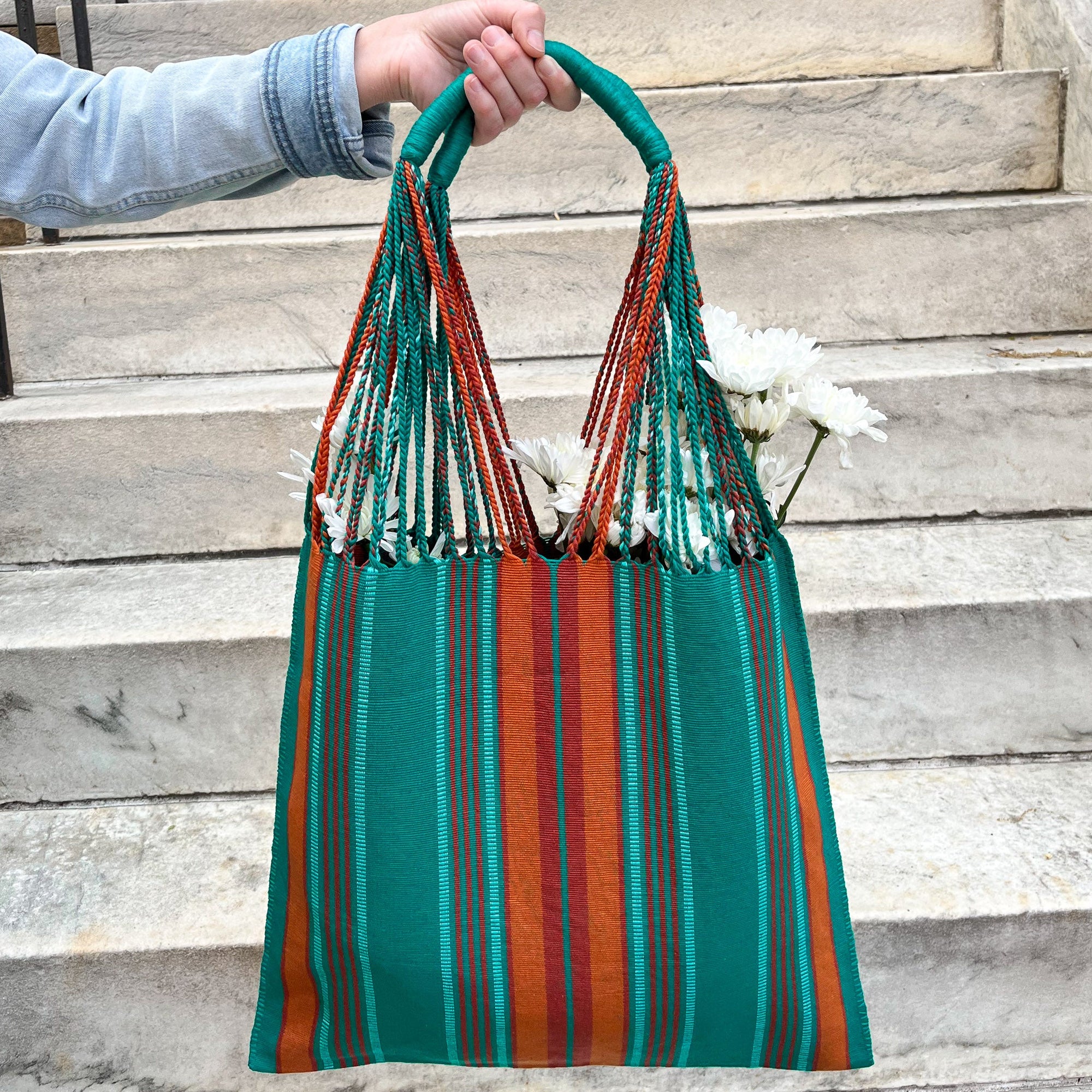 Vibrant handwoven teal and orange market bag, made in Guatemala, held in an outstretched hand against a backdrop of white stone stairs. The bag is filled with fresh-cut white flowers