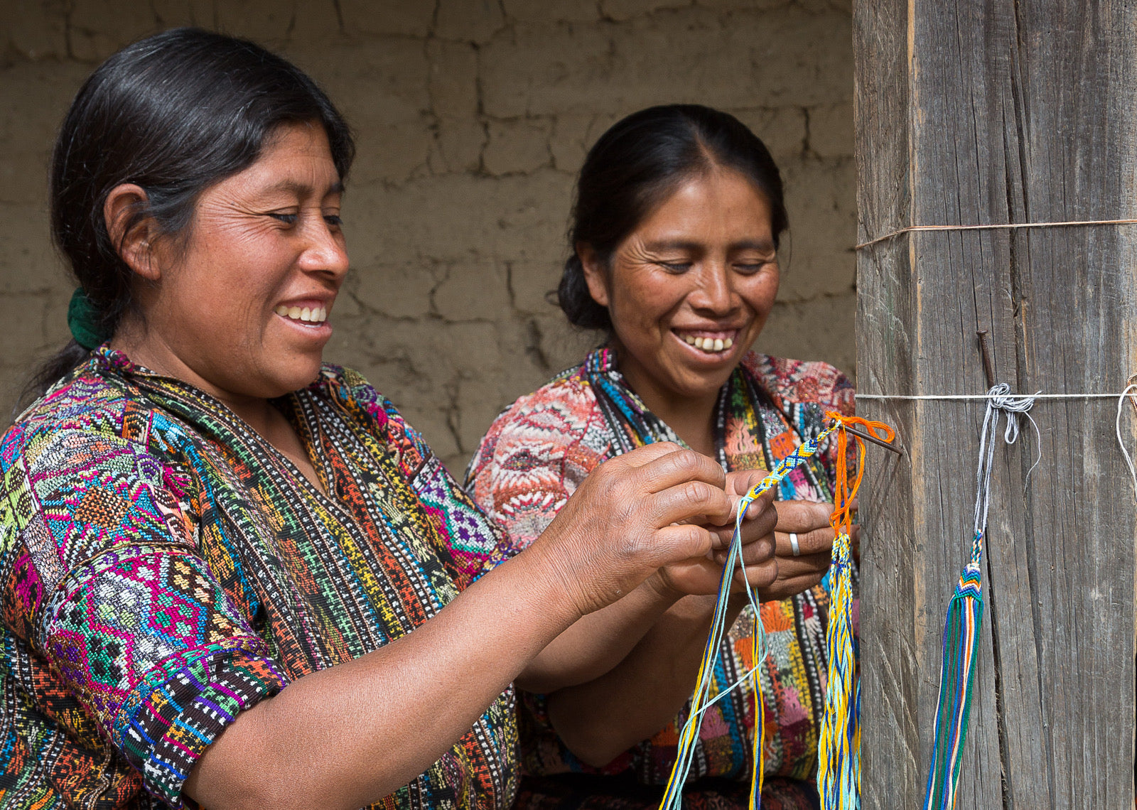 Two smiling adult women pictured from the waist up weave friendship bracelets from colorful thread, with the bracelets attached to a wooden pole. They wear handwoven huipiles from Sololá Guatemala and appear to be laughing.