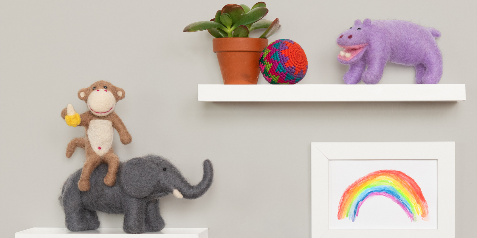 Whimsical needle felted animal figures of an elephant, monkey holding a banana, and purple hippo sit on white shelves mounted on a light grey wall with a small plant and a framed rainbow artwork
