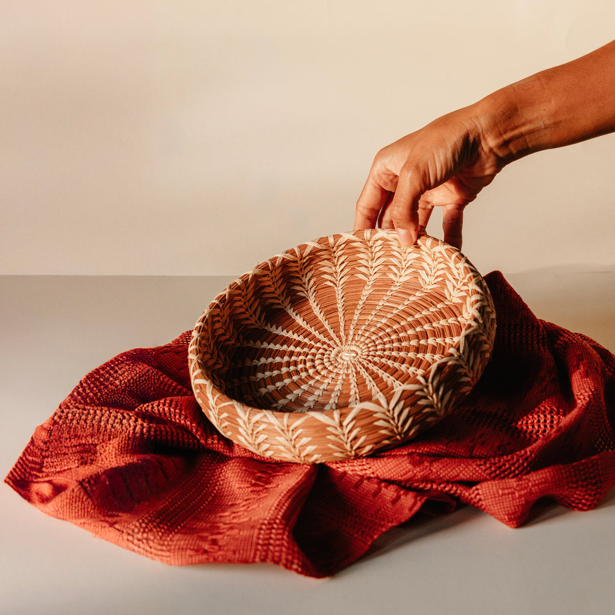 Hand holding Cecilia Basket, with basket resting on draped terracotta table runner in front of soft white background.