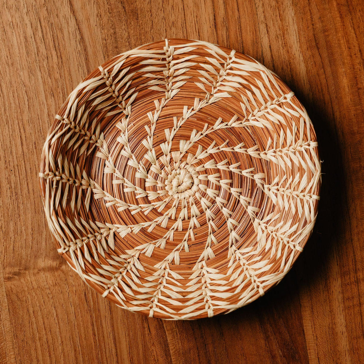 A round woven basket with a spiral pattern, resting on a warm brown wooden surface.