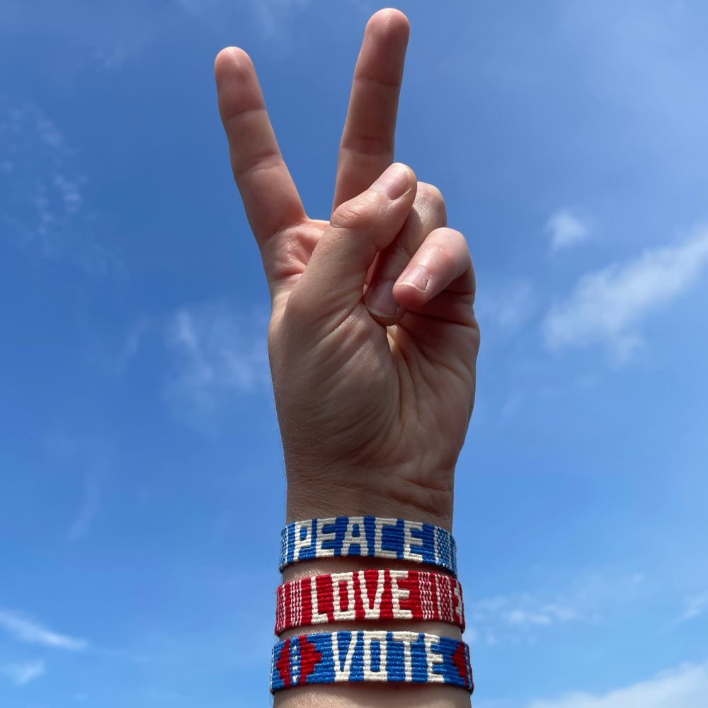 A hand making a peace sign against a blue sky, wearing three woven fabric wristbands that read 'PEACE', 'LOVE', and 'VOTE' in red, white, and blue.
