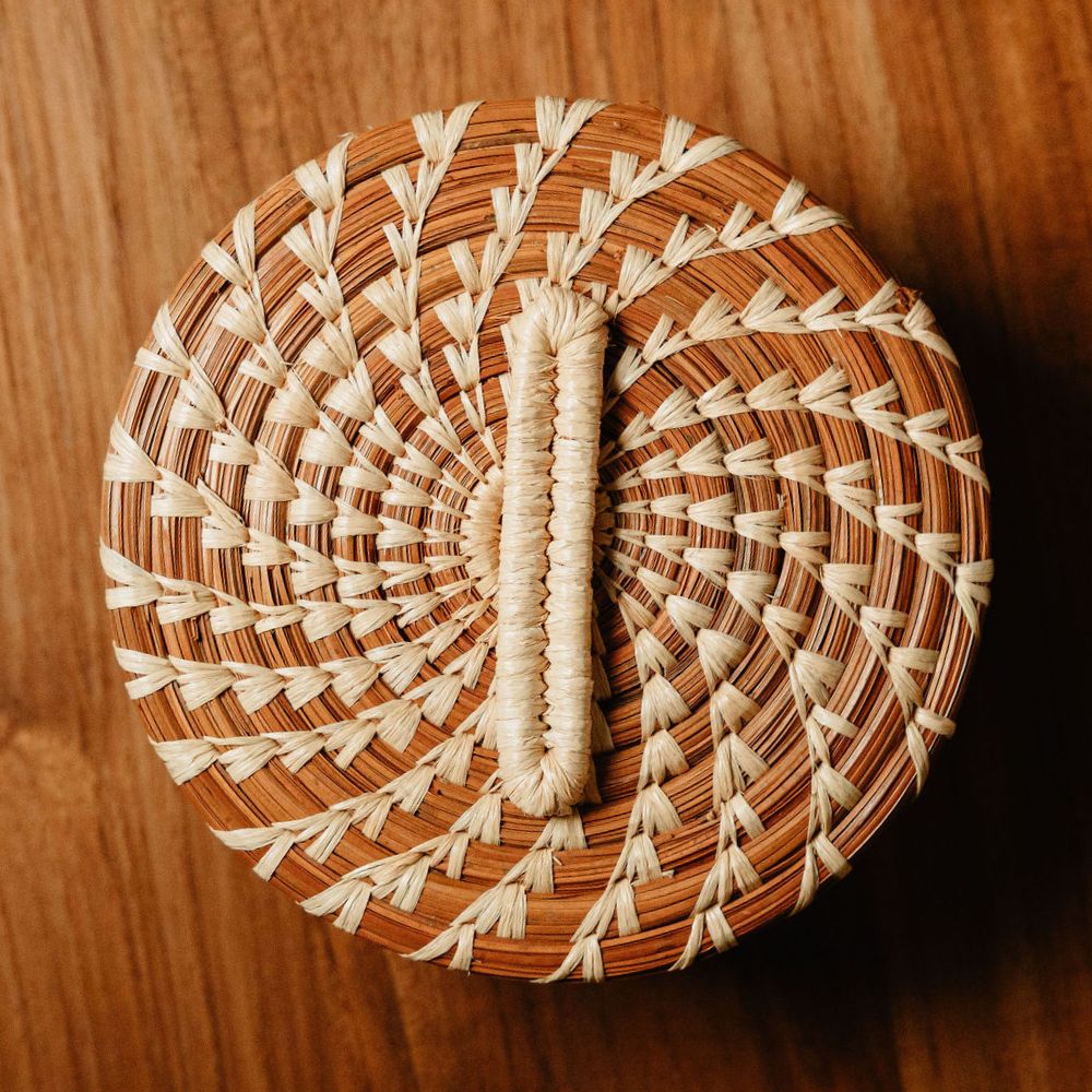 A round woven basket lid with a central handle, photographed from above on a warm wooden surface. The lid features a natural brown base with intricate light beige and cream woven patterns radiating outward.