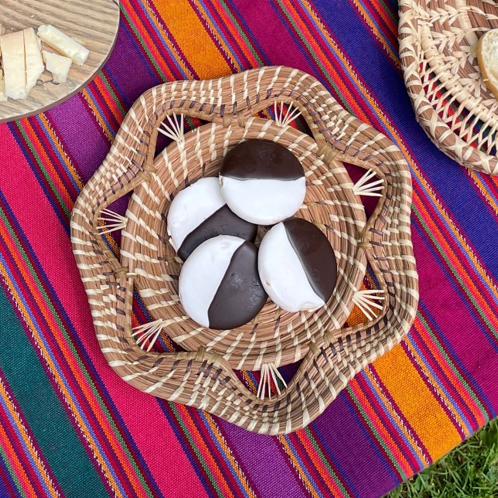 Four chocolate-dipped cookies arranged in a woven pine needle basket on a colorful striped textile, with partial wooden and basket trays visible at the top left and right.