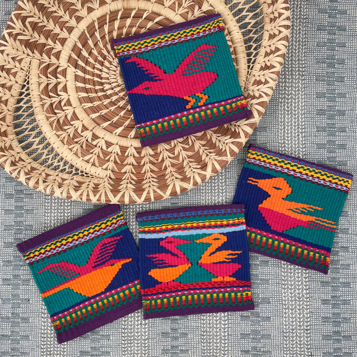 Set of four colorful handwoven pajarito coasters displayed over Calado table runner and Angela pine needle basket tray with mesh detail on handles.